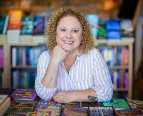 Andrea Guevara, author brand strategist leaning on stacks of books at a bookstore
